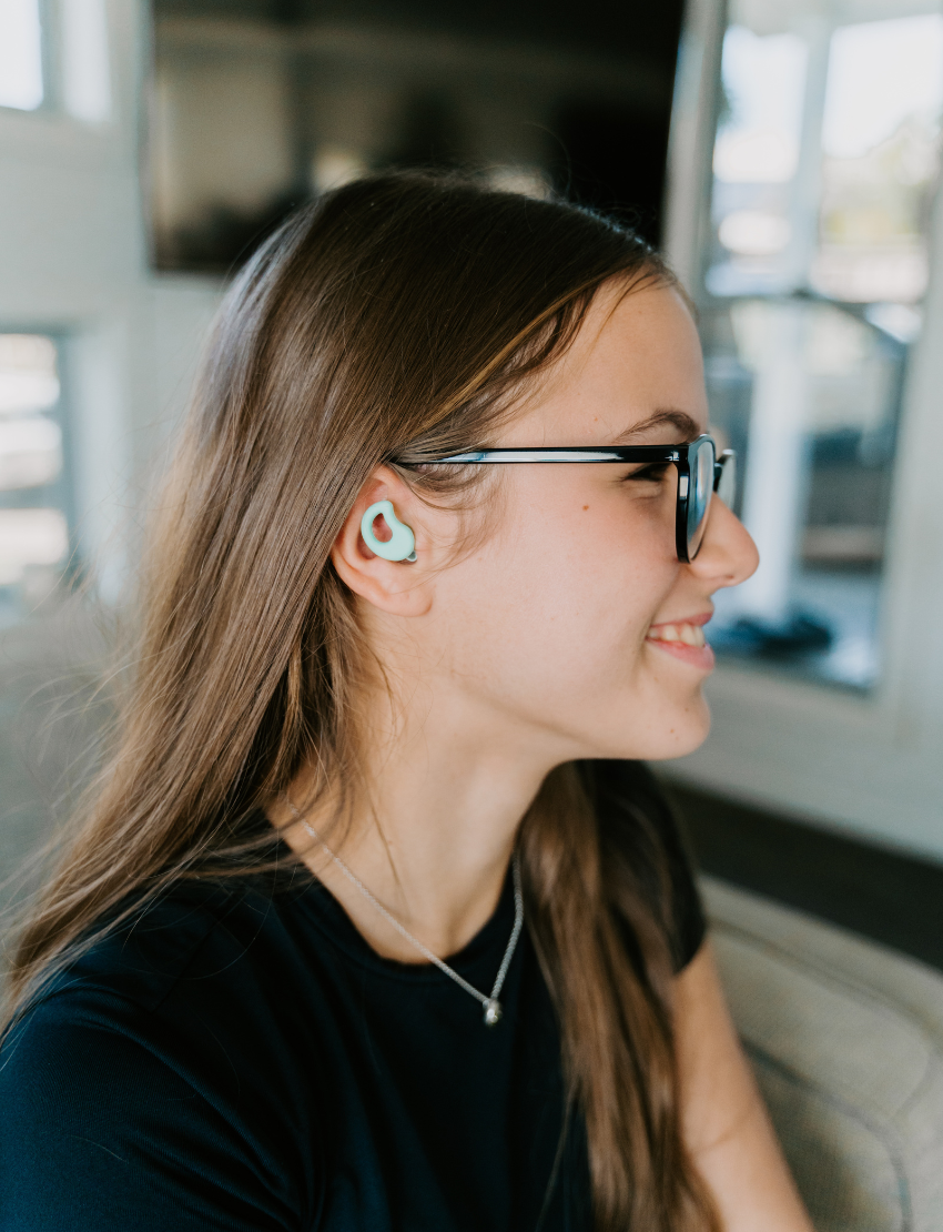 Teenage girl with glasses wearing a teal Curvd Earplug from The Play Way, smiling, experiencing improved sensory focus.