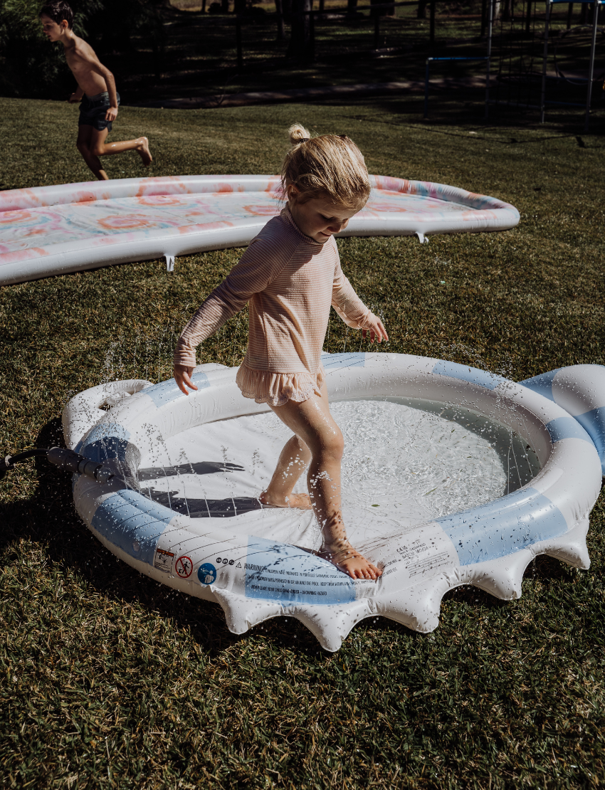 A child in a pink swimsuit plays in a blue and white SunnyLife Sprinkler Mat by The Play Way, promoting sensory regulation.