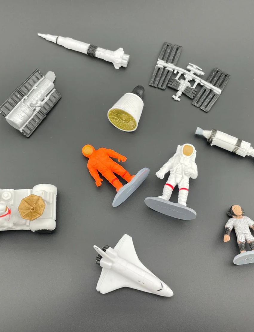 Collection of space-themed miniature figures including astronauts, rockets, and the ISS on a dark background.