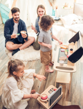A family playing with a wooden BBQ grill play set, with children and parents interacting.