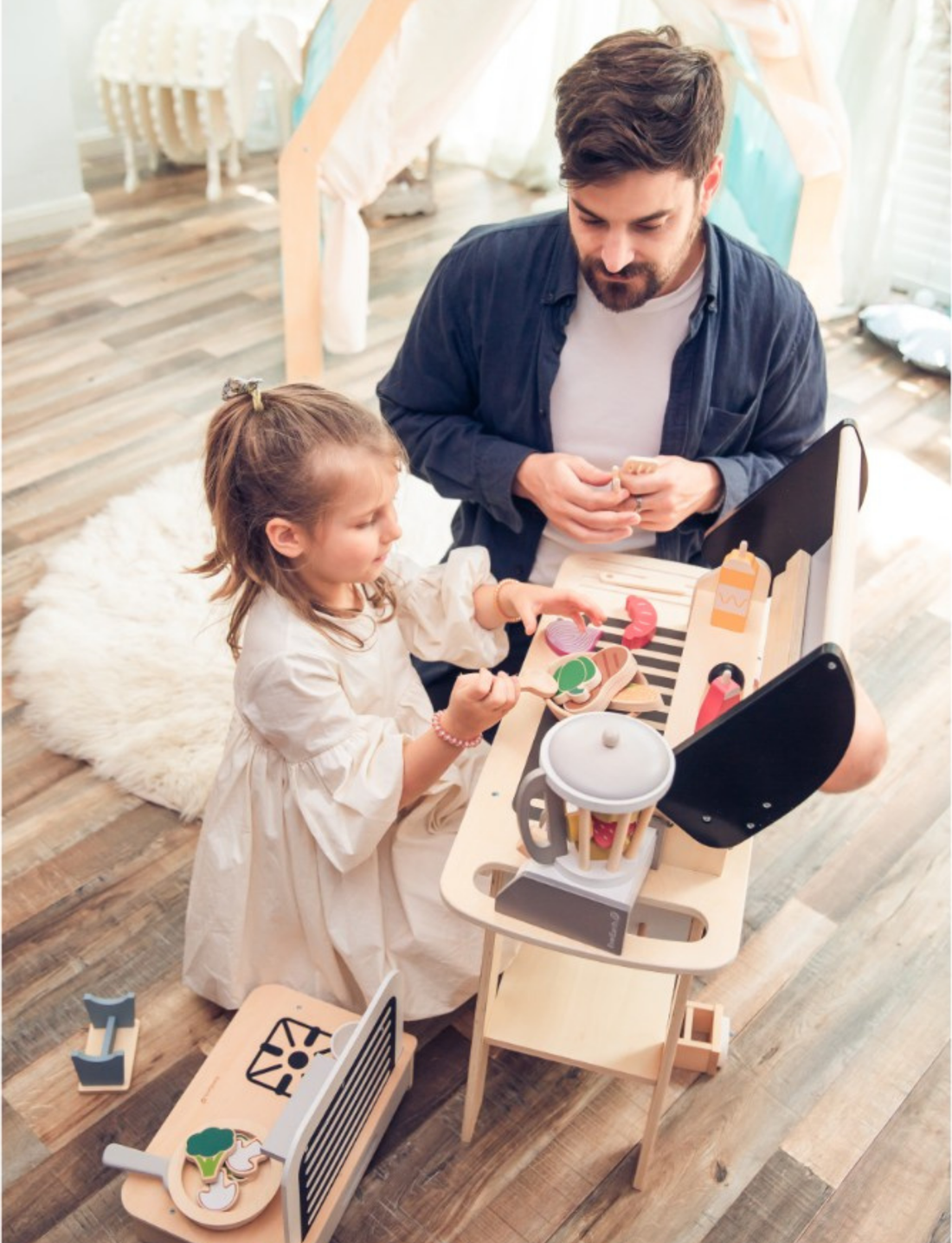 A wooden BBQ play set with a grill, wheels, and accessories like ketchup and mustard bottles, on a white background.