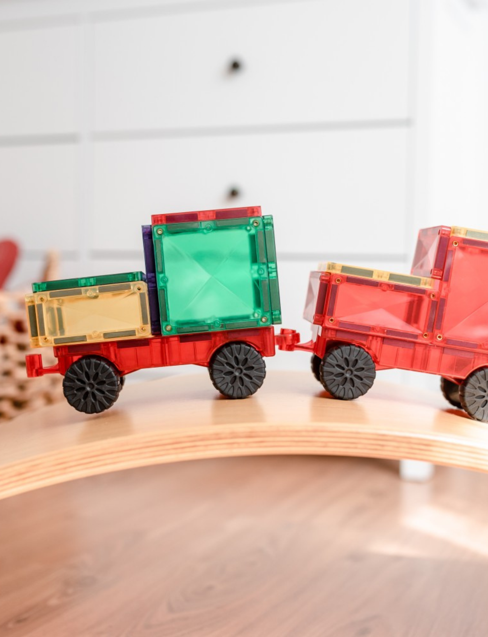 Two Connetix magnetic tile train cars, one red with black wheels, carrying colorful magnetic tiles, on a wooden surface.
