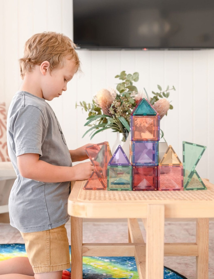 Boy building with Connetix pastel magnetic tiles, creating a colorful house and tower on a wooden table.