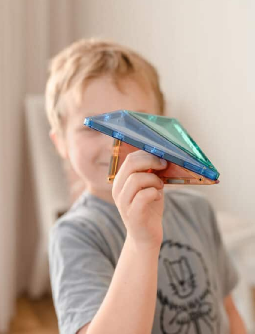Young boy holding and playing with translucent blue and green Connetix magnetic tiles, forming a plane.