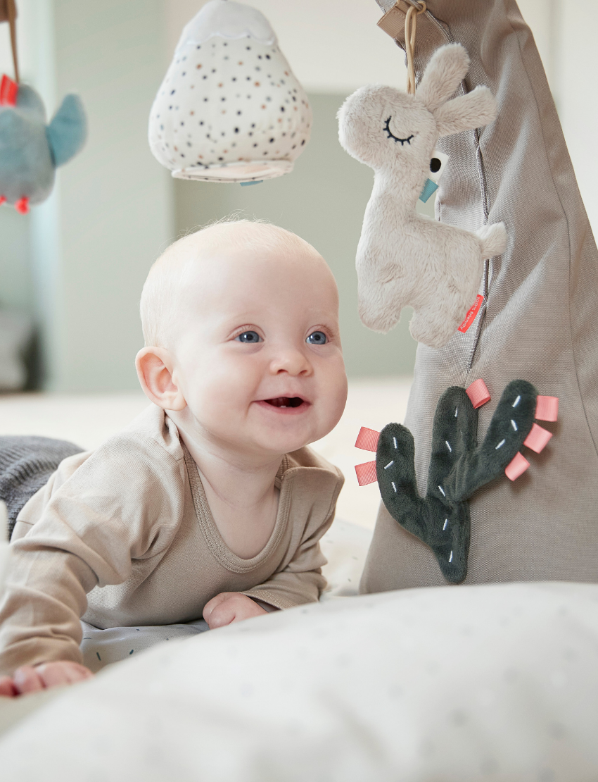 Smiling baby lying under a grey activity gym with plush llama, bird, and cactus toys