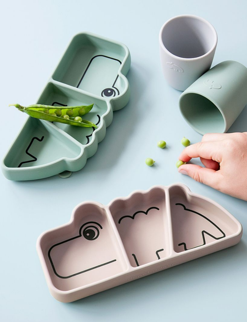 Sage green and pink silicone crocodile plates on a blue surface, with a hand adding peas to the green plate.