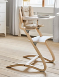 Wooden high chair with light beige cushions and a white detachable tray, set in a room.