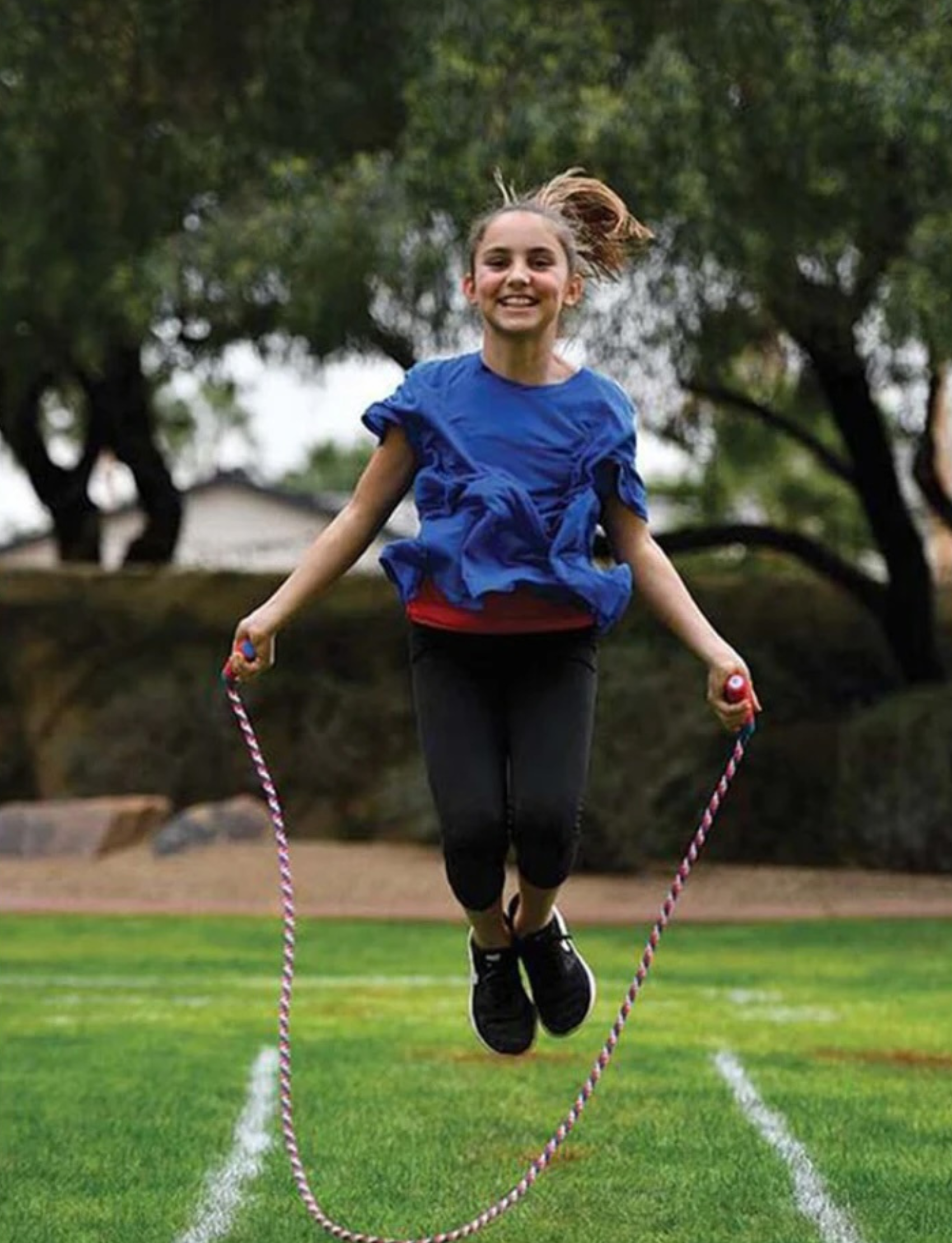 A smiling girl in mid-air, jump roping in an outdoor obstacle course with agility lines marked on the grass.
