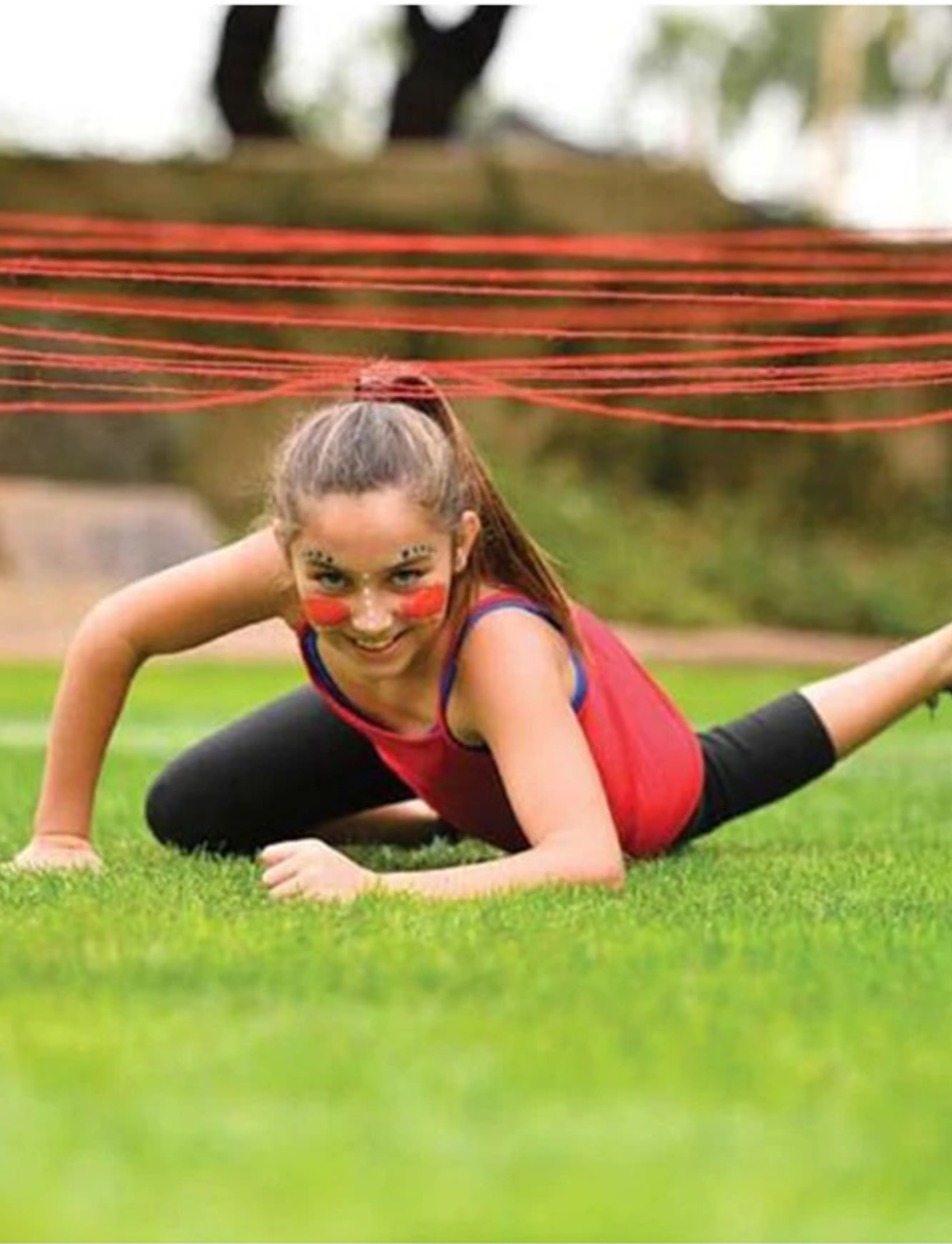 A young girl crawling under a red string obstacle in an outdoor agility course, face painted with 'ninja' markings.