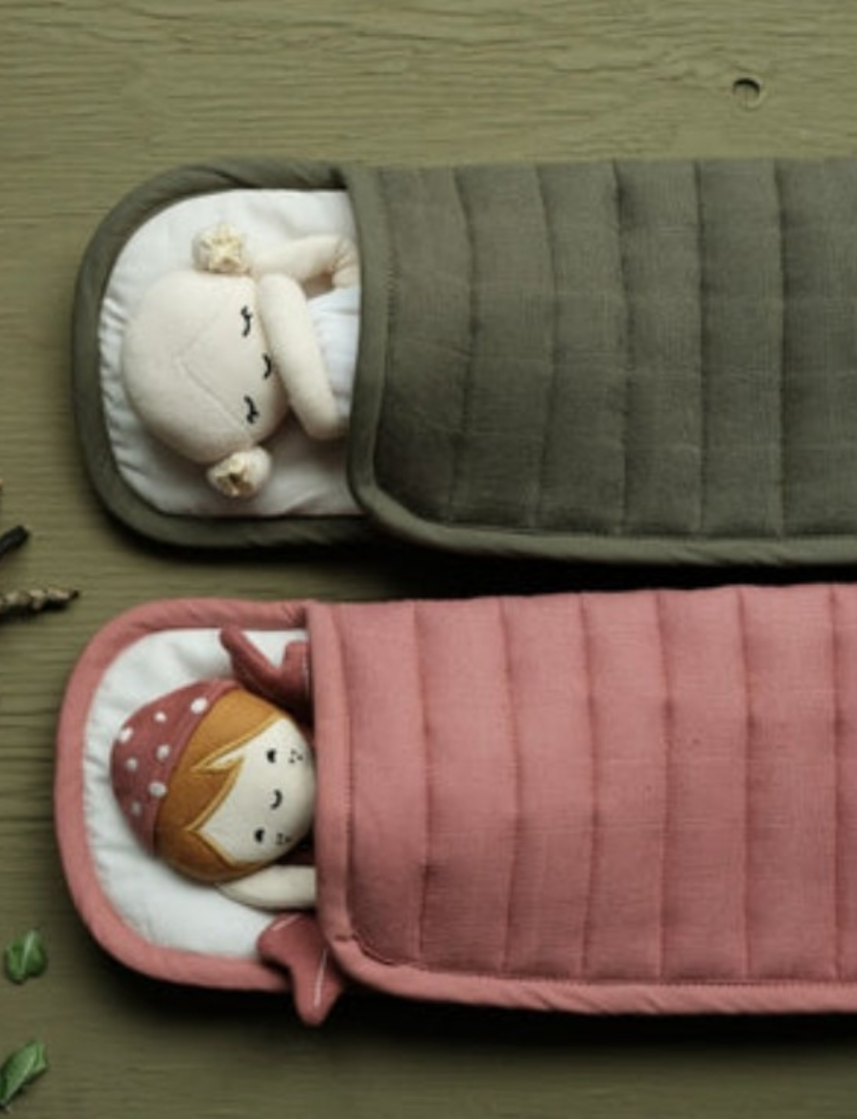 Two fabric dolls lying in sleeping bags, one green and one pink, within a camping setting.