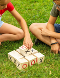 Children's hands placing wooden blocks on grass for outdoor Tic Tac Toe game.