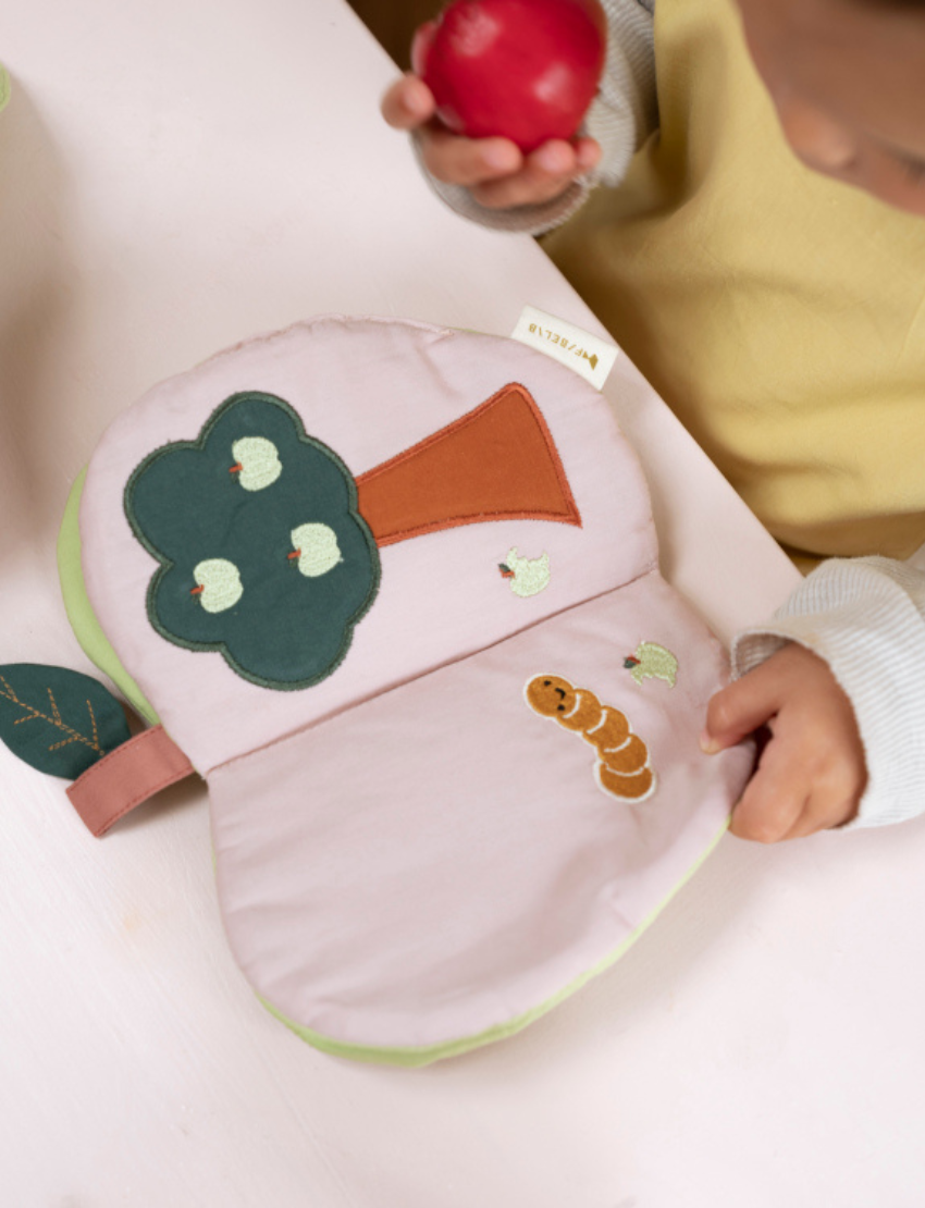 Child holding a red apple and looking at an open fabric book with a tree and worm applique.