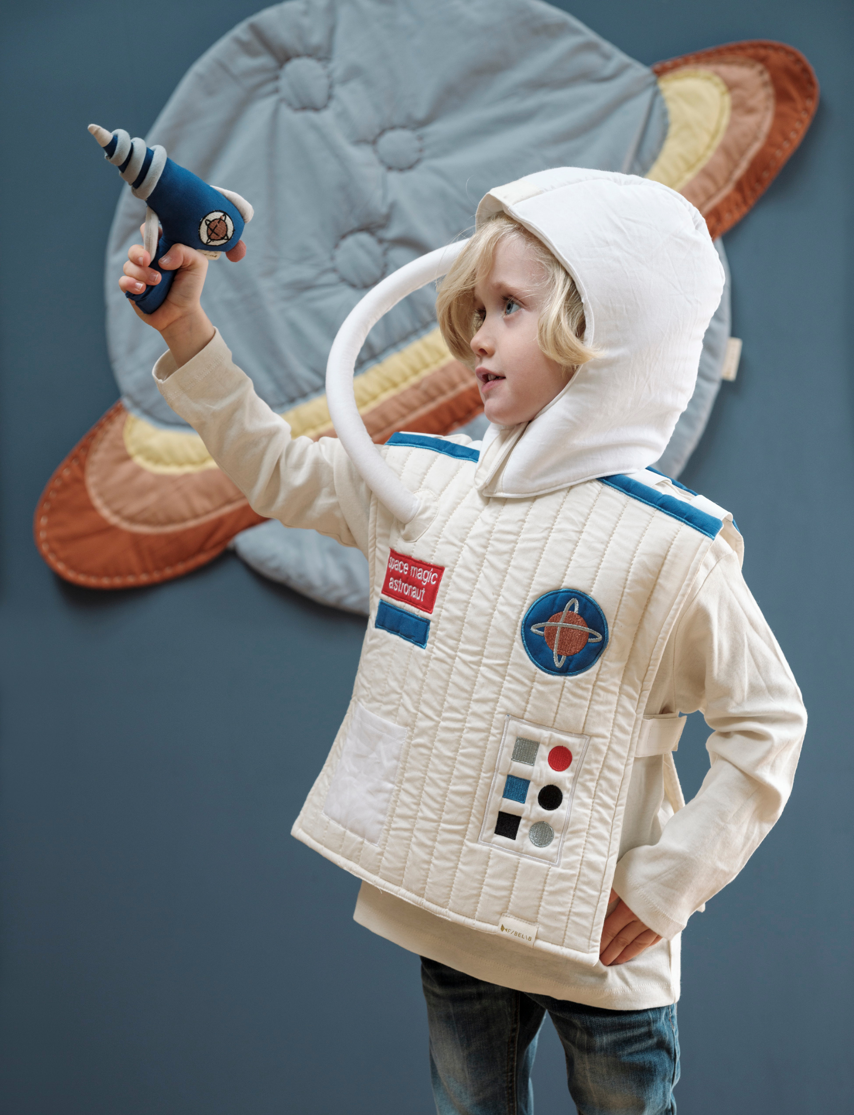 Young child in a white astronaut costume with a helmet, holding a blue toy ray gun, with a planet decoration behind.