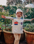 Happy child in an astronaut costume, wearing pink glasses, standing outdoors in pots with plants.
