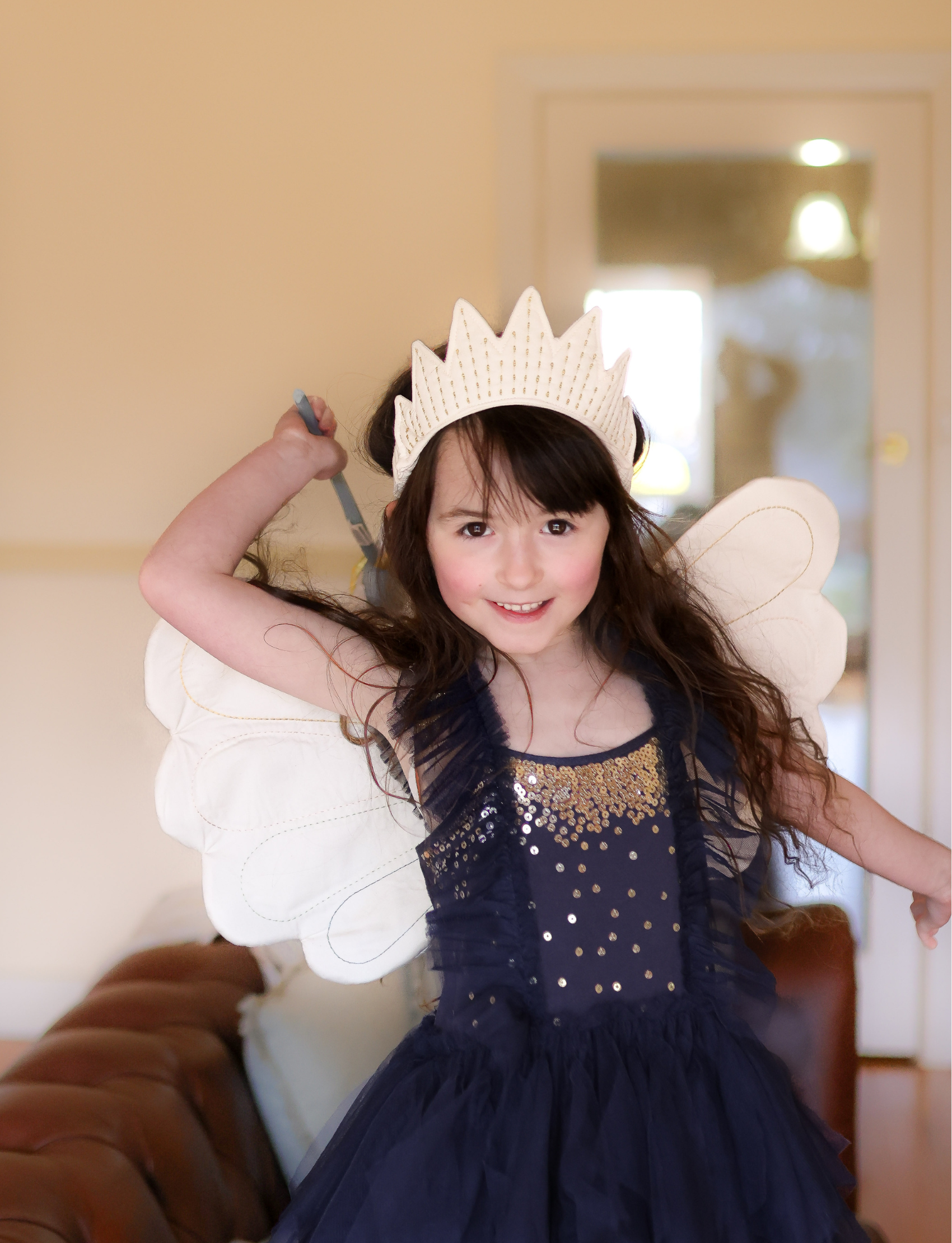 A little girl dressed as a fairy with a crown, wings, and dark blue sparkly dress, holding a wand.