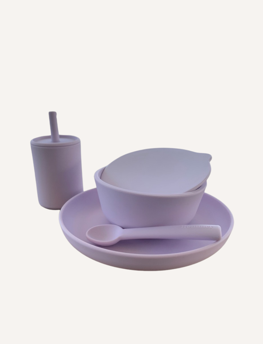 Lilac baby dinner set featuring a cup with a straw lid, a bowl with a lid, and a spoon on a matching plate.