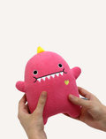 Pink plush dinosaur toy held by hands, featuring cute embroidered face and yellow spikes.