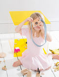 A young child in a pink dress sits on a white floor, playing with a wooden toy camera and other toys.