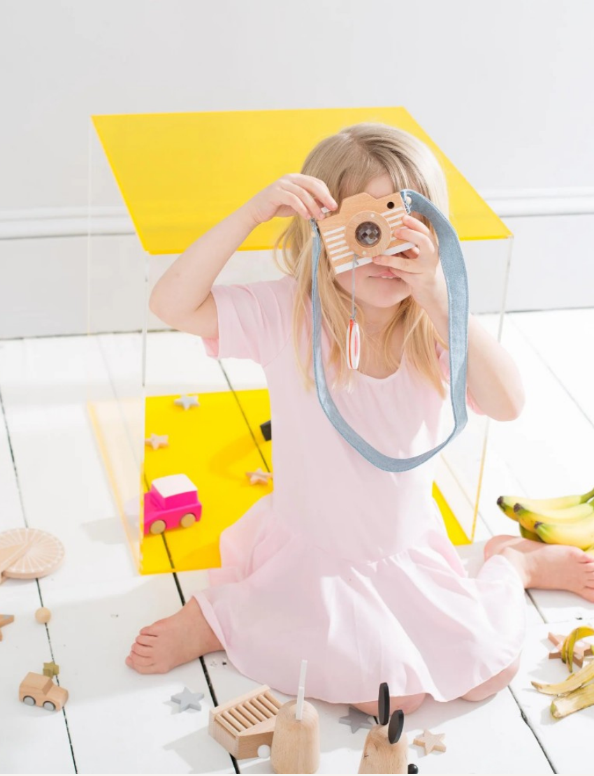 A young child in a pink dress sits on a white floor, playing with a wooden toy camera and other toys.