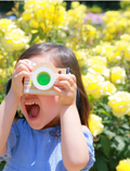 A little girl with dark hair wears a blue shirt and holds a wooden toy camera with a green lens to her face.