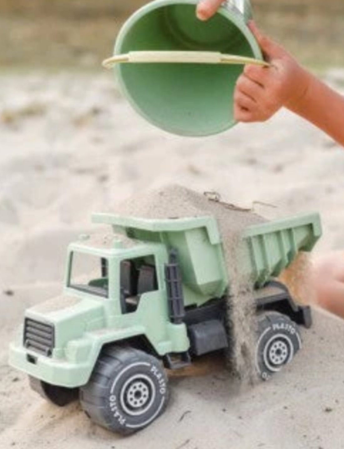 Green toy sandpit set with tipper truck, bucket, scoop, and sand mold, against a pale background.