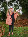 A young girl dressed as a ladybug, wearing a cape and headband, pointing outwards in a garden setting.