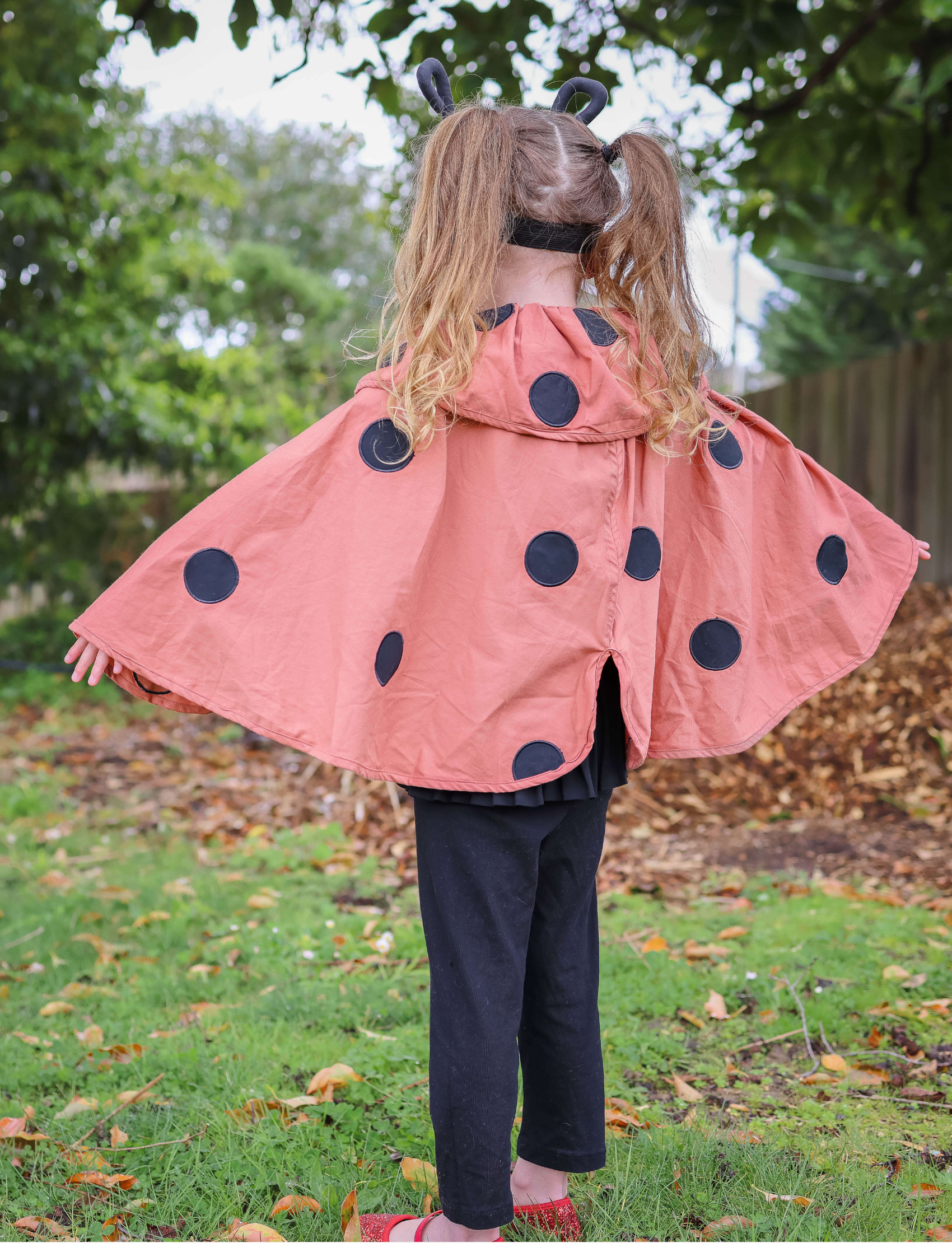 Back view of a young girl in a ladybug costume with a polka dot cape spread out, standing in a garden.