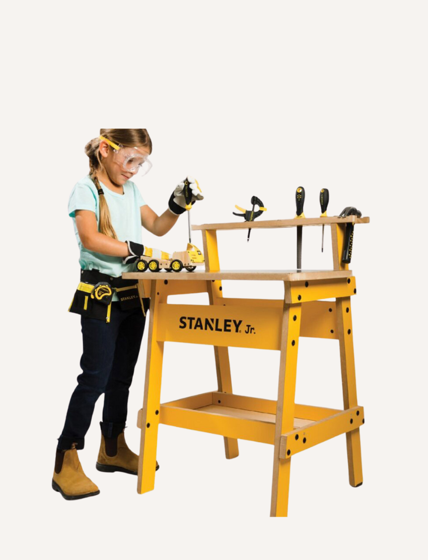 Girl in safety goggles and tool belt works at a yellow Stanley Jr. kids' workbench with toy tools.