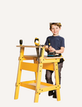 Smiling boy wearing goggles and tool belt uses a saw at a yellow Stanley Jr. kids' workbench.