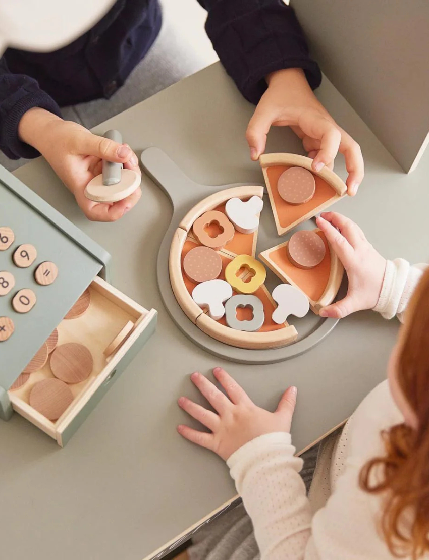 Wooden pretend play pizza set featuring a sage green pizza peel, multi-colored sliceable toppings, and a matching wooden pizza cutter.
