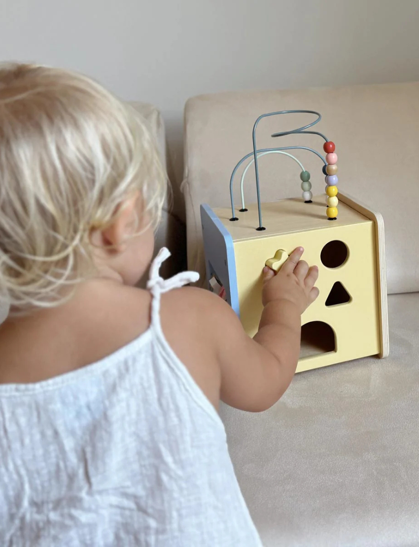 Moover wooden activity cube with shape sorting holes on a yellow side and a cute cat face on a striped blue side.