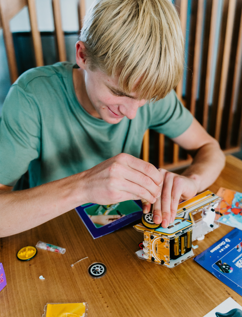 Young man with blonde hair assembling the CircuitMess Sparkly robot car, focusing on attaching a wheel.