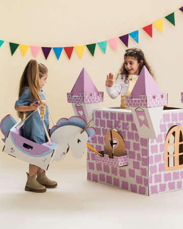 Two children playing with a pink cardboard castle and unicorn carriage, one child waving from castle.