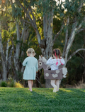 Two young girls walking in a grassy field with one wearing a cardboard horse costume.