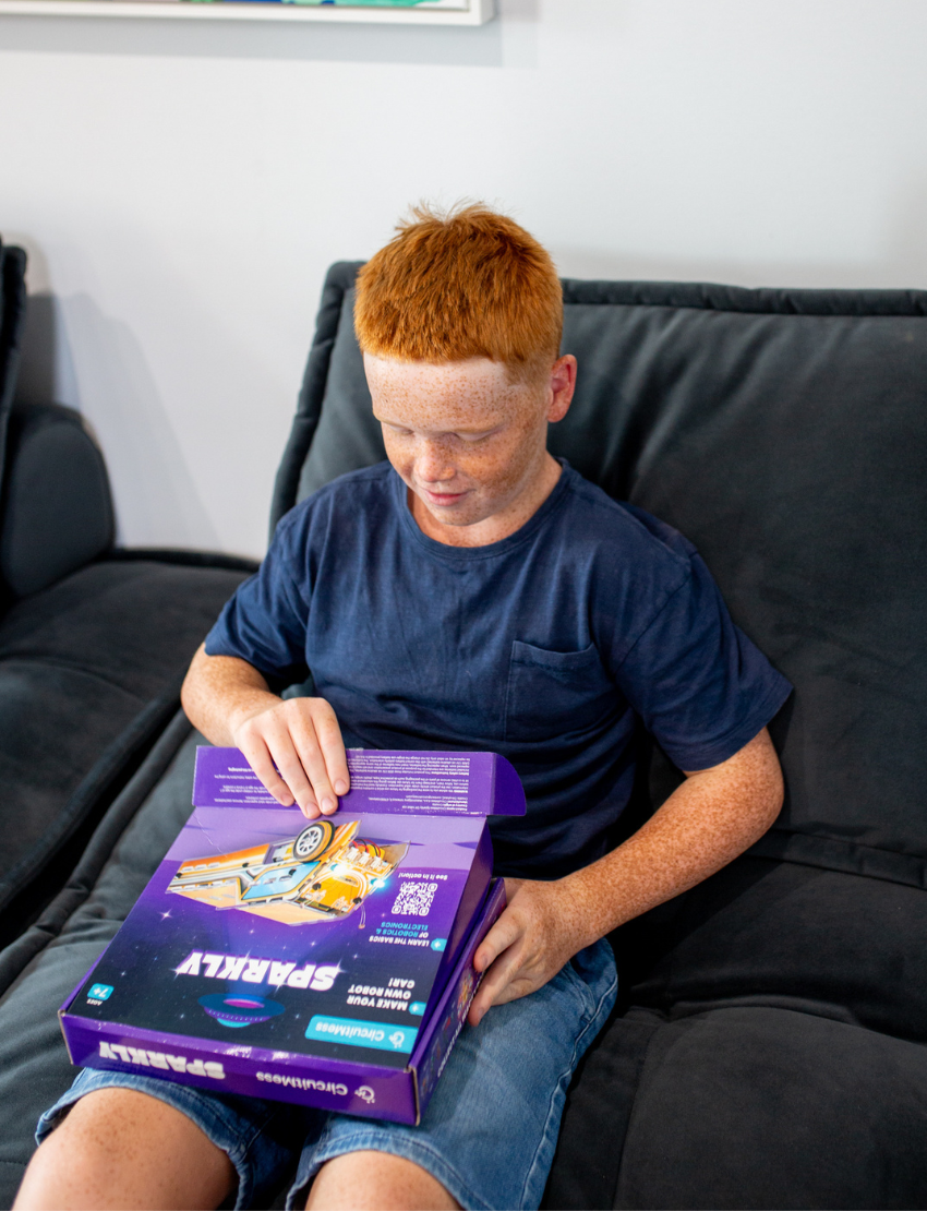 Young boy with red hair and freckles opening the CircuitMess Sparkly robot car kit box.