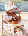 Baby in a fruit-patterned bucket hat and portable high chair, with silicone feeding set, for convenient mealtimes.