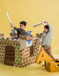 Two boys in a cardboard castle, one with a sword, the other with a cardboard horse, engaged in imaginative play.