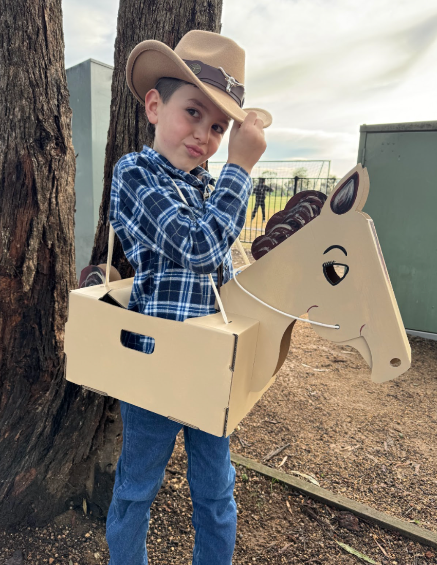 Boy dressed as a cowboy wears a cardboard horse costume, posing outdoors.