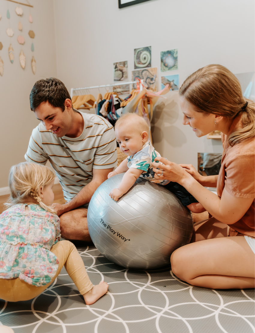 The Tummy Time Program by Therapy At Home comes in an open teal box with abstract shapes and includes a silver exercise ball, bubble wand, baby food pouch, cards, leaflets, and packaging for baby development and parent guides.