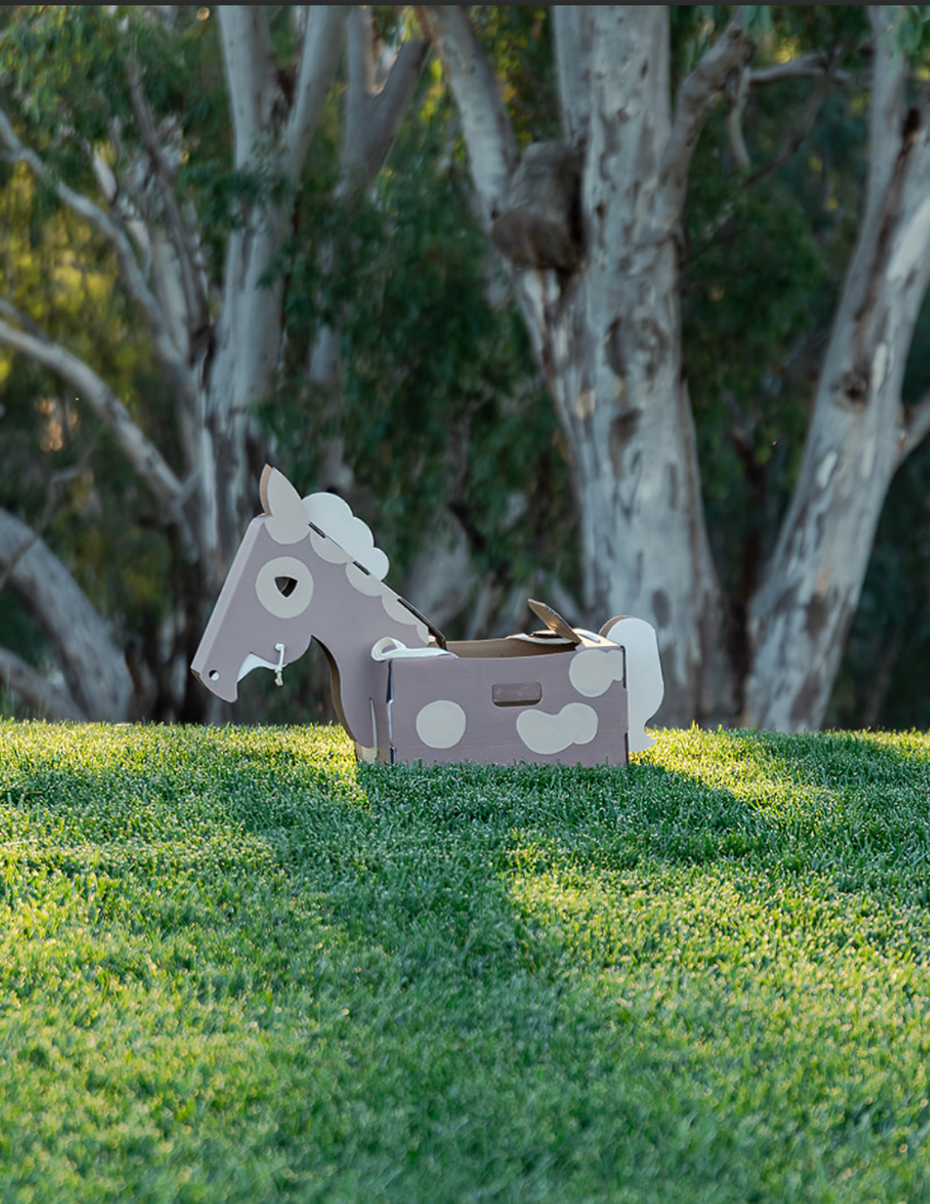A grey and white cardboard horse toy with polka dots sits on a green grassy hill.