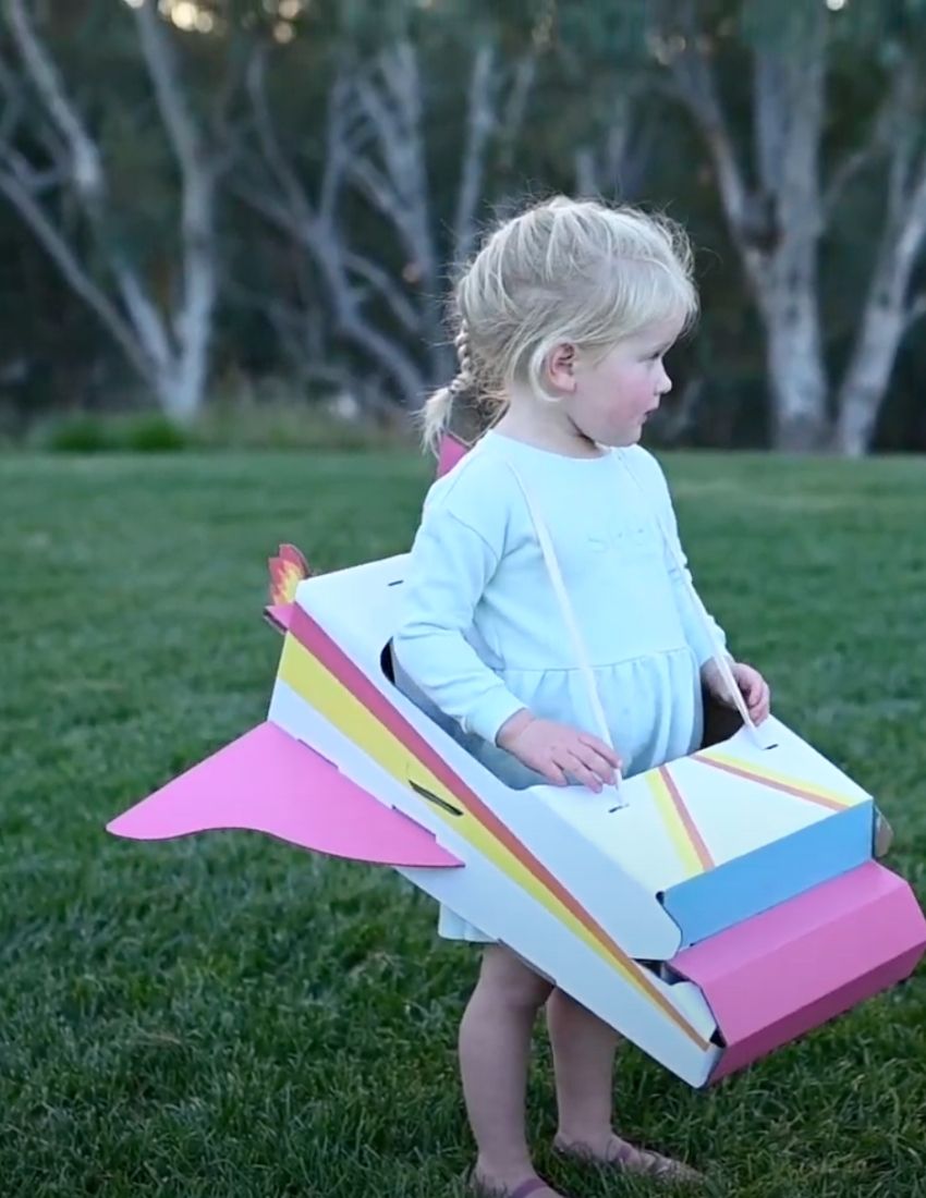 Child wearing a colorful cardboard airplane costume, playing outdoors.