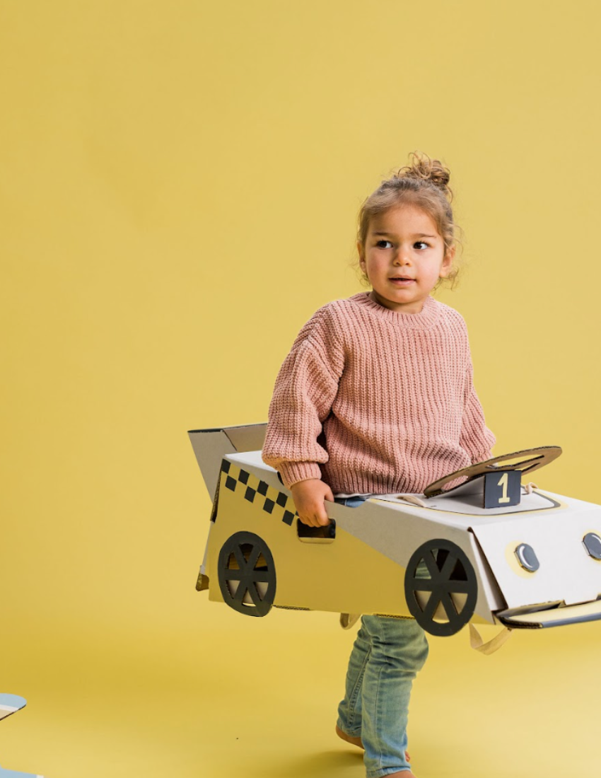 Young girl wearing a pink sweater holding a cardboard race car costume.