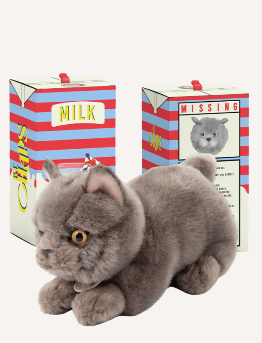 Grey plush cat wearing a party hat, sitting in front of stylized 'MILK' and 'MISSING' packaging boxes.