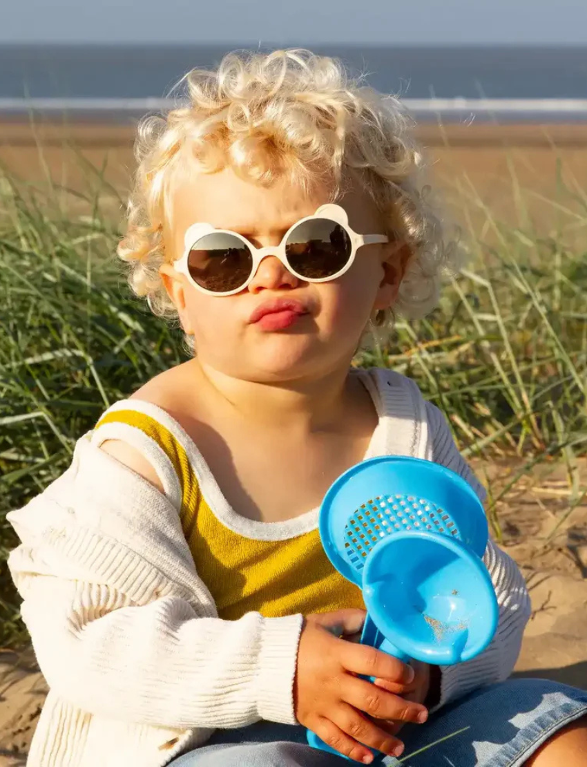Cute toddler with curly hair wearing cream bear-shaped sunglasses and a yellow top on the beach.