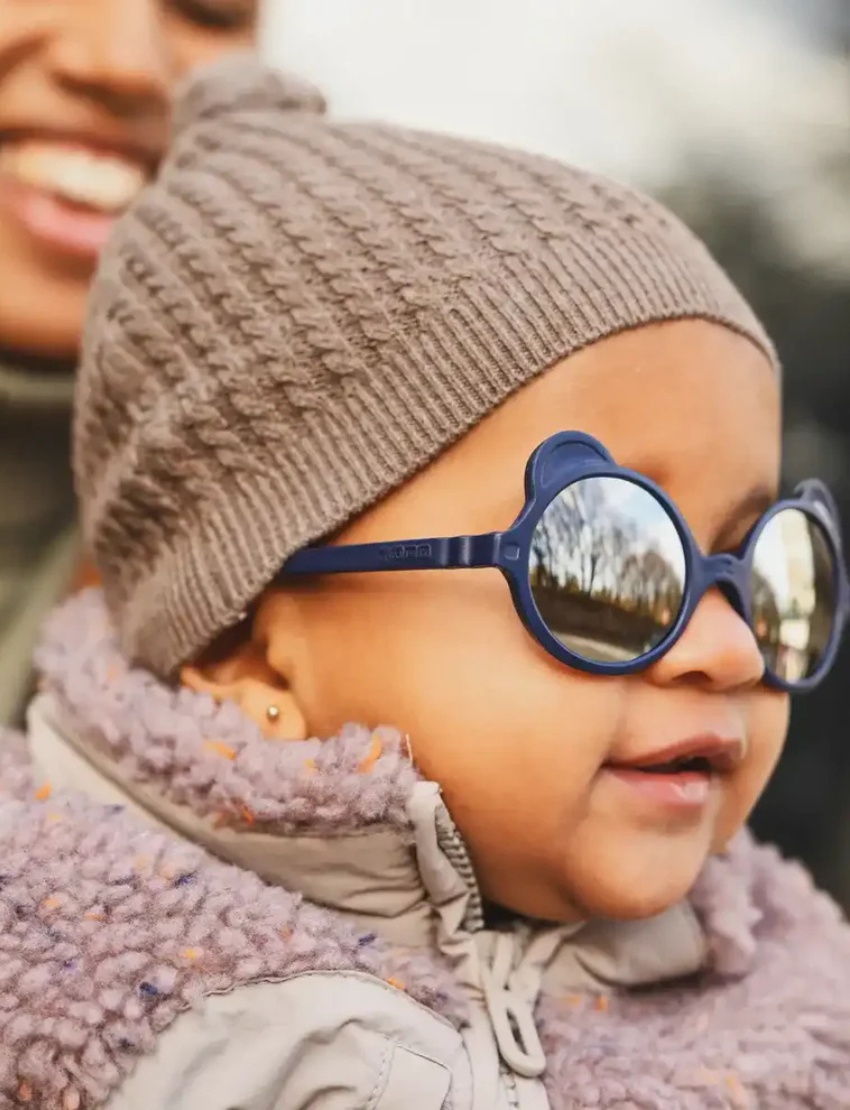 Adorable baby wearing navy blue bear-shaped sunglasses and a knitted hat.