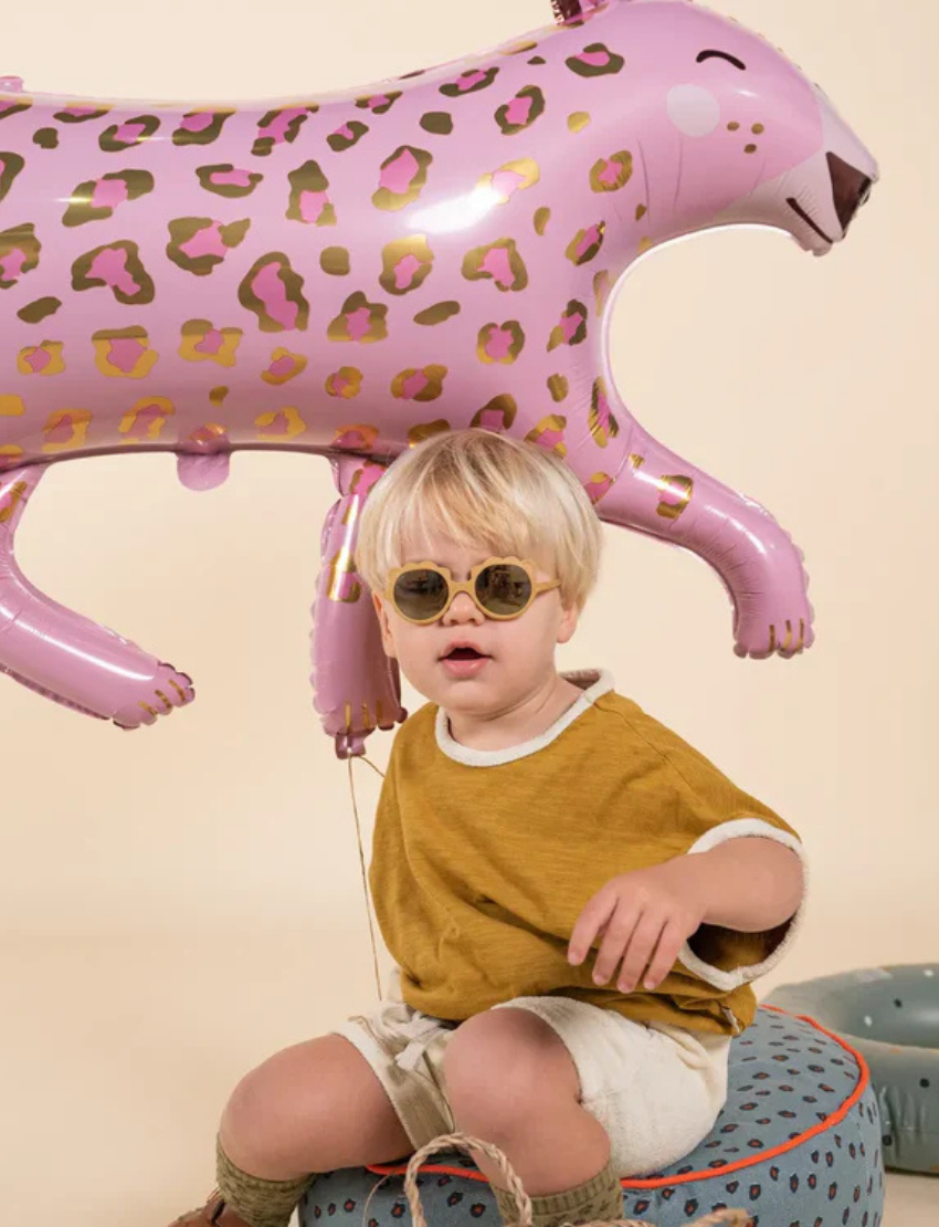 Young boy wearing round tan Ki ET LA sunglasses and a mustard shirt, sitting in front of a pink leopard balloon.