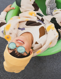 Smiling child wearing teal Ki ET LA sunglasses and a yellow beanie, lying in a green tube.