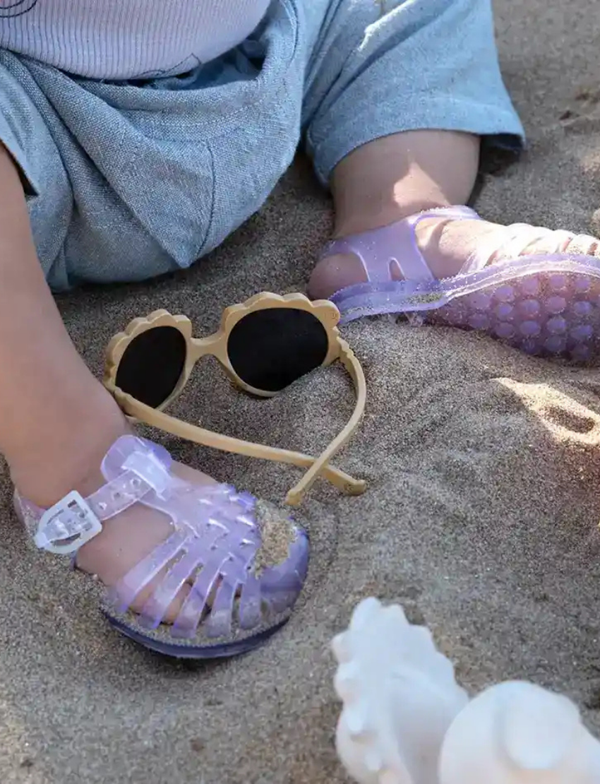Close-up of a toddler's feet wearing sparkly purple jelly sandals and tan Ki ET LA sunglasses in the sand.