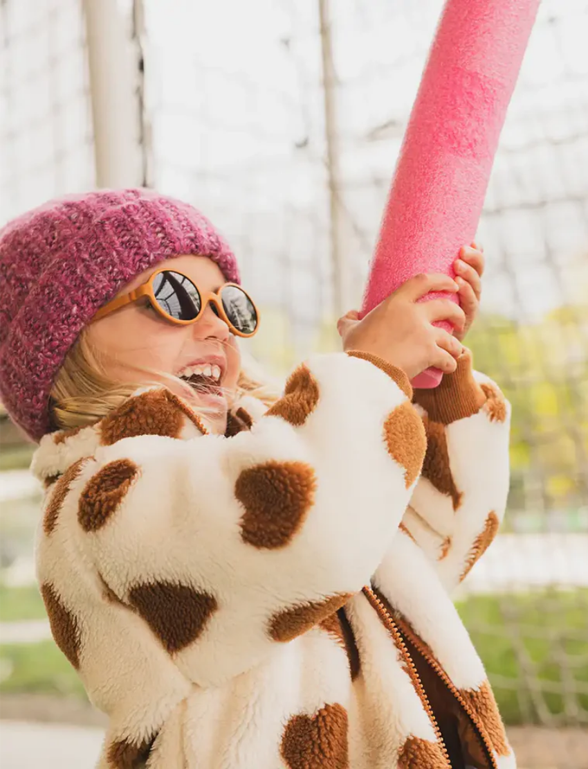 Happy young girl in a pink beanie, cow-print jacket, and sunglasses, playfully holding a pink foam noodle outdoors.