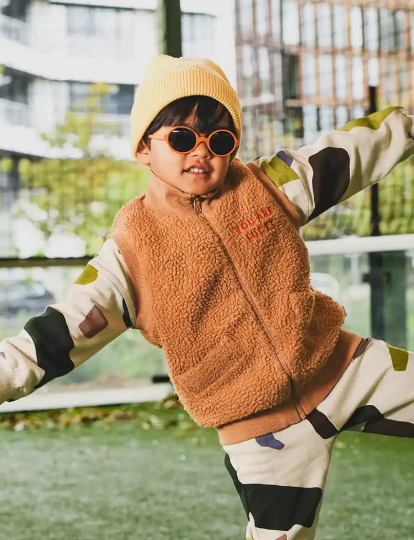 Happy boy in yellow beanie, orange round sunglasses, brown fleece vest, and camo sweatsuit, perfect for kids' outdoor style.